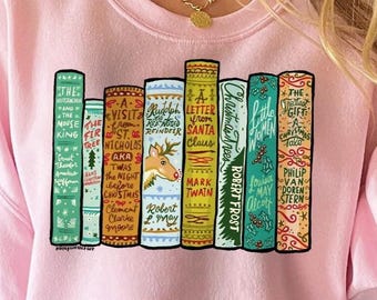 May include: A light pink sweatshirt featuring a colorful graphic of stacked books. The book spines display titles like "Visit St. Nicholas" and "Christmas Clues." The sweatshirt is paired with blue jeans.