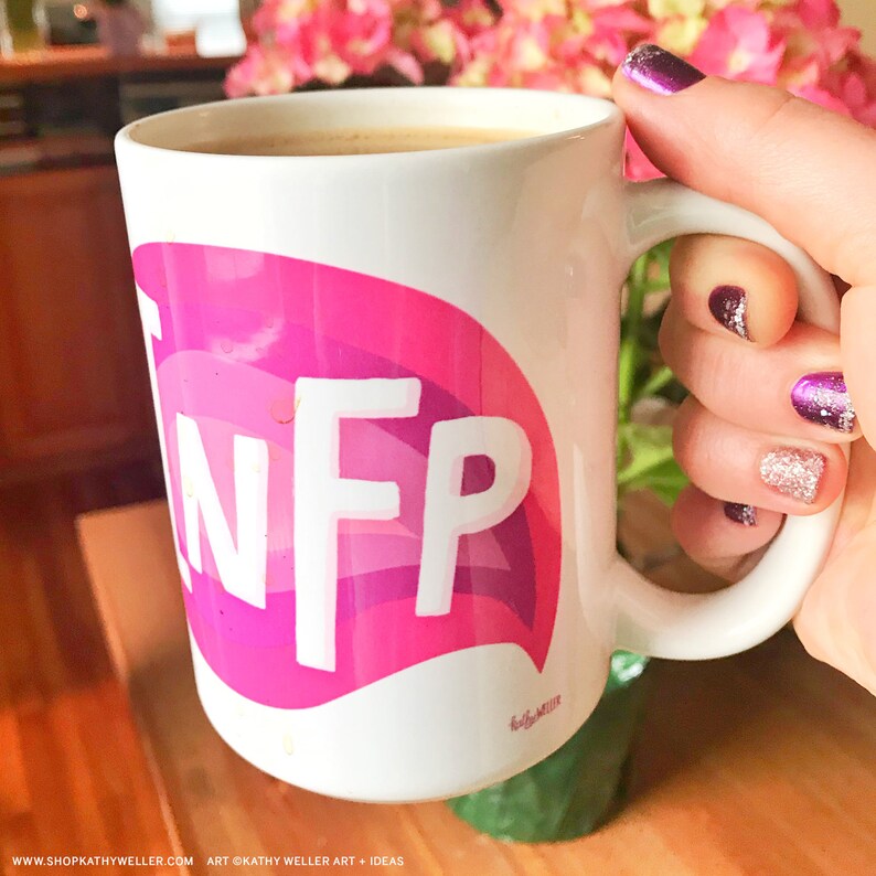 May include: White ceramic mug with a pink and purple speech bubble design that says "INFP". The mug is filled with coffee and is being held by a hand with purple nail polish.