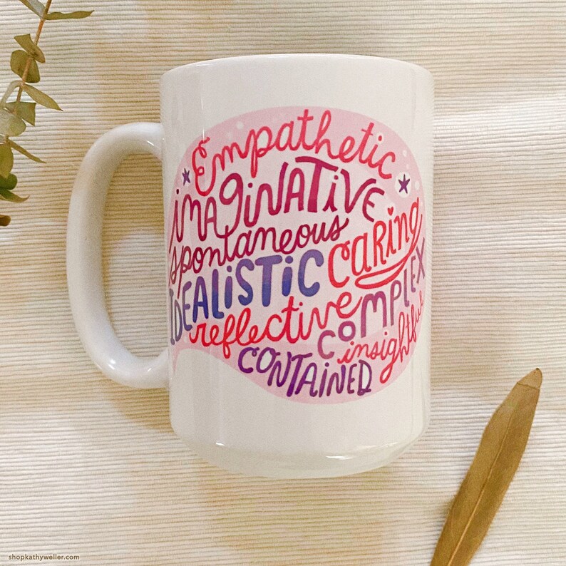 May include: White ceramic mug with a pink and purple design. The design features a brain shape with the words "Empathetic", "Imaginative", "Spontaneous", "Caring", "Realistic", "Reflective", "Complex", "Contained", and "Insightful" written inside.