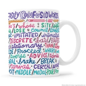 May include: White ceramic mug with a colorful design of words that are easily confused, such as 'lose' and 'loose', 'council' and 'counsel', 'brake' and 'break'. The words are written in a handwritten style with different colors.