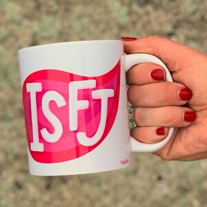 May include: White ceramic mug with a pink and red design featuring the letters "ISFJ" in white. The mug has a handle and is being held by a hand.