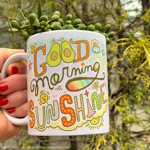 May include: White ceramic mug with a colorful hand-drawn design that says "Good Morning Sunshine". The design includes avocados, suns, and rainbows.