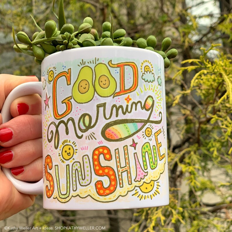 May include: White ceramic mug with a colorful hand-drawn design that says "Good Morning Sunshine". The design includes avocados, suns, and rainbows.