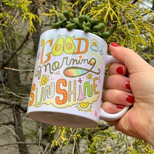 May include: White ceramic mug with a colorful hand-drawn design that says "Good morning sunshine". The design includes avocados, suns, rainbows, and stars. The mug is being held by a hand with red nail polish.