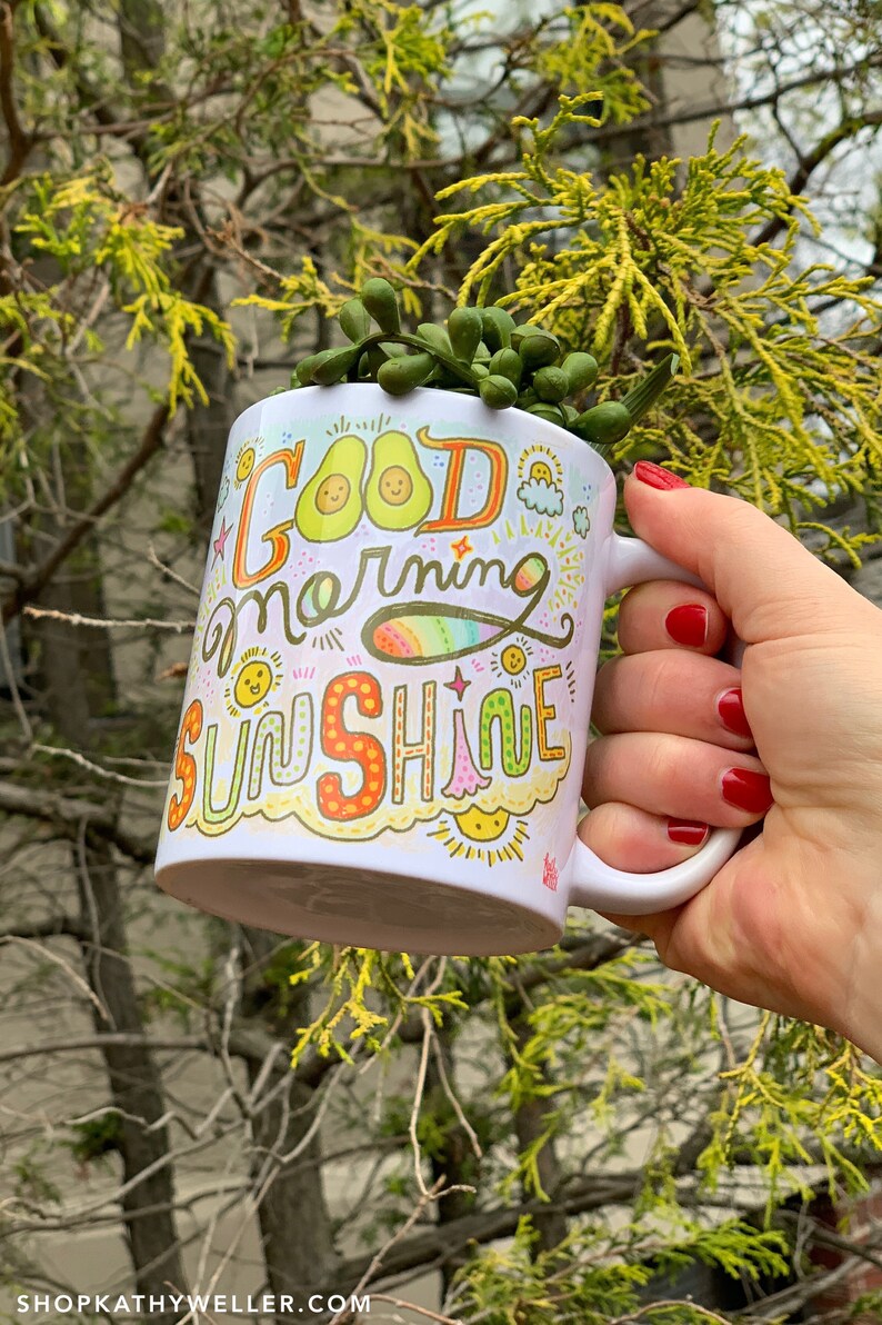 May include: White ceramic mug with a colorful hand-drawn design that says "Good morning sunshine". The design includes avocados, suns, rainbows, and stars. The mug is being held by a hand with red nail polish.