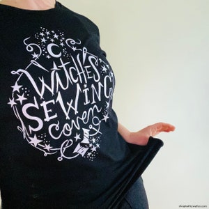 May include: Black t-shirt with a white graphic design that says "Witches Sewing Coven" with stars and sewing-related imagery.