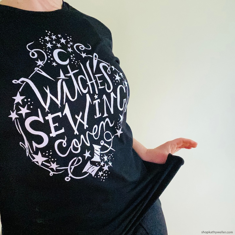 May include: Black t-shirt with a white graphic design that says "Witches Sewing Coven" with stars and sewing-related imagery.