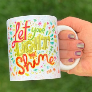 Puede incluir: Taza de cer&aacute;mica blanca con las palabras "Let Your Light Shine" en letras coloridas y caprichosas. La taza tiene un asa blanca y est&aacute; decorada con varios dise&ntilde;os coloridos de estrellas y flores.