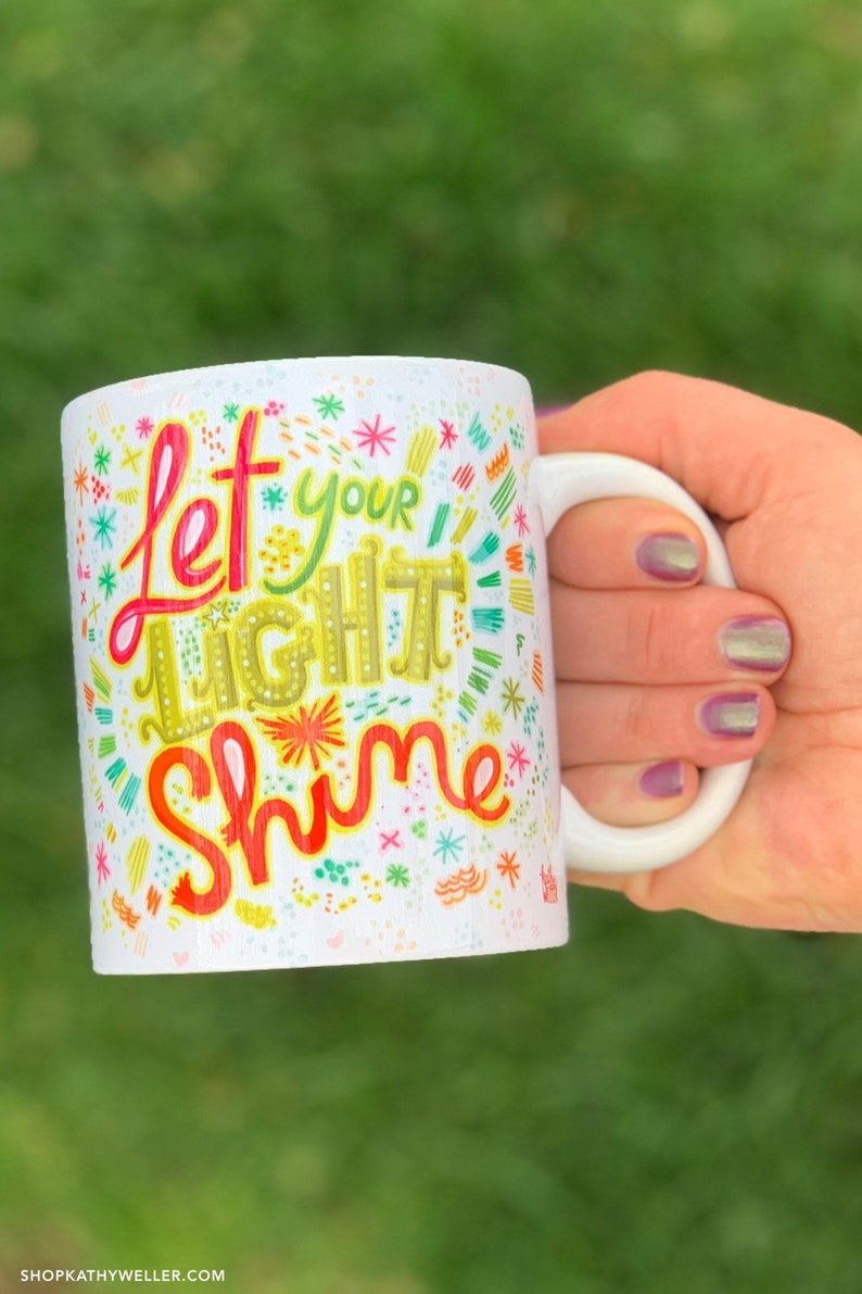 Puede incluir: Taza de cer&aacute;mica blanca con las palabras "Let Your Light Shine" en letras coloridas y caprichosas. La taza tiene un asa blanca y est&aacute; decorada con varios dise&ntilde;os coloridos de estrellas y flores.