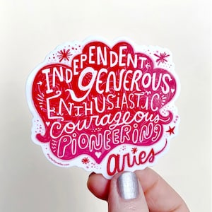 May include: A red and white sticker featuring the zodiac sign Aries and a list of personality traits: independent, generous, enthusiastic, courageous, pioneering. The sticker is being held in a hand with silver nail polish.