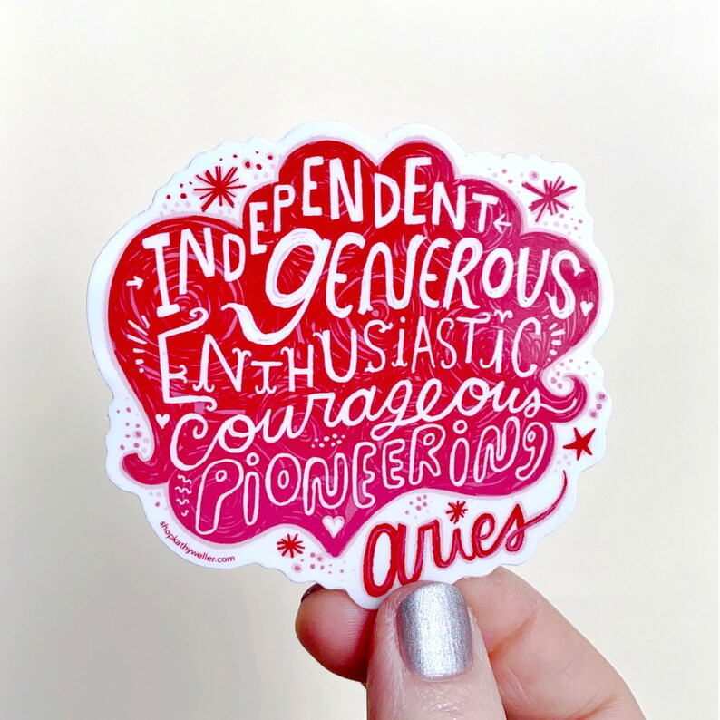 May include: A red and white sticker featuring the zodiac sign Aries and a list of personality traits: independent, generous, enthusiastic, courageous, pioneering. The sticker is being held in a hand with silver nail polish.