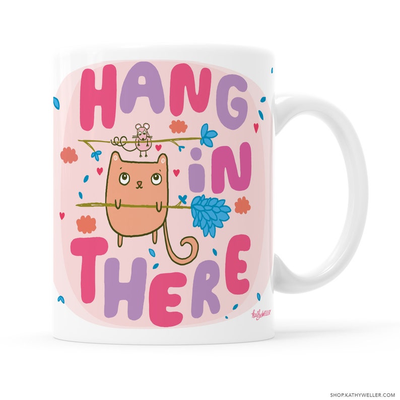 May include: White ceramic mug with a pink background and a cartoon cat holding a branch with a mouse on it. The text "Hang in there" is written in pink, purple, and blue letters.
