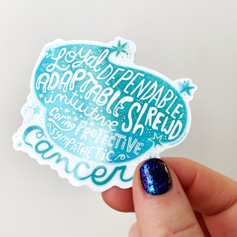 May include: A teal and white sticker with the word "Cancer" and related personality traits like "Loyal" and "Adaptable" in a handwritten font. The sticker has a watercolor effect and star accents.