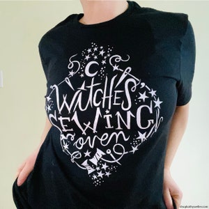 May include: Black t-shirt with a white graphic design that says "Witches Brewing Coven" with stars and other witchy symbols.
