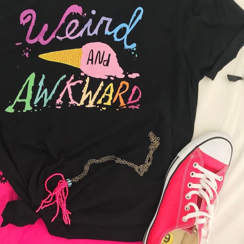 May include: Black t-shirt with the text "Weird and Awkward" in rainbow colors. The text is stylized to look like an ice cream cone. A pink and gold necklace is draped over the shirt. A pink and white sneaker is in the lower right corner of the image.
