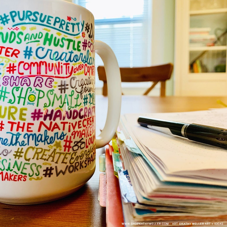 May include: White ceramic mug with colourful text that reads "#PURSUE PRETTY #HUSTLE #CREATOR #COMMUNITY #SHARE #SHOPSMALL #HANDCRAFT #NATIVE #THE MAKERS #CREATE #BUSINESS #WORKSPACE". The mug is sitting on a wooden table next to a stack of papers and a black pen.