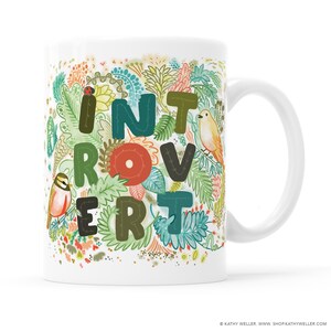May include: White ceramic mug with a colorful floral design and the word "Introvert" spelled out in large, stylized letters. The letters are outlined in black and have a stitched appearance.