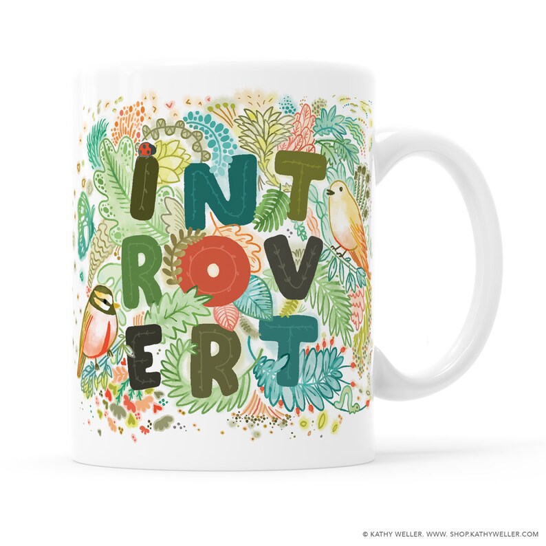May include: White ceramic mug with a colorful floral design and the word "Introvert" spelled out in large, stylized letters. The letters are outlined in black and have a stitched appearance.