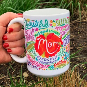May include: White ceramic mug with a colorful design featuring the word "Mom" surrounded by positive words like "beautiful", "supportive", "fiercely", "loving", and "teacher".