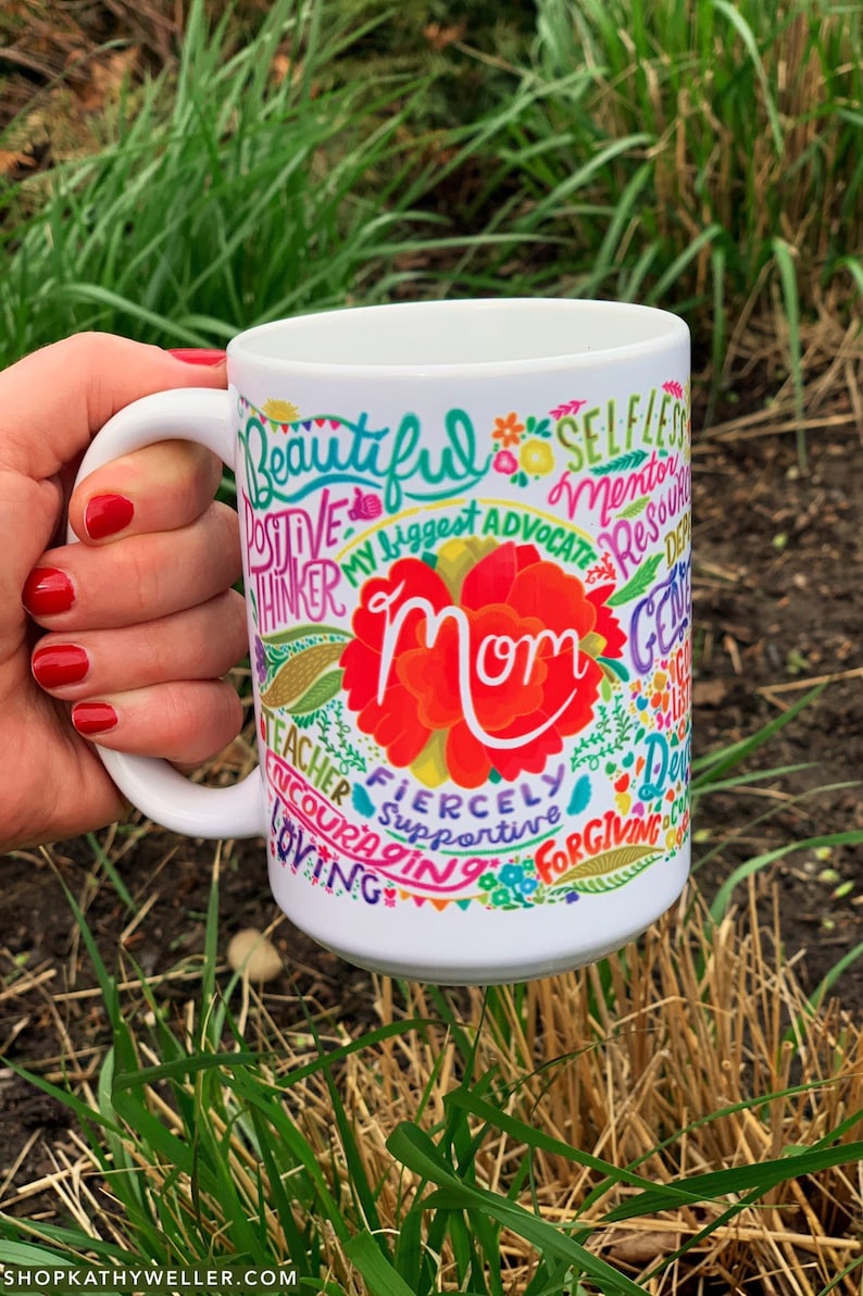 May include: White ceramic mug with a colorful design featuring the word "Mom" surrounded by positive words like "beautiful", "supportive", "fiercely", "loving", and "teacher".