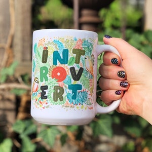 May include: White ceramic mug with a colorful floral design and the word "Introvert" written in a whimsical font. The mug is being held by a hand with black nail polish.