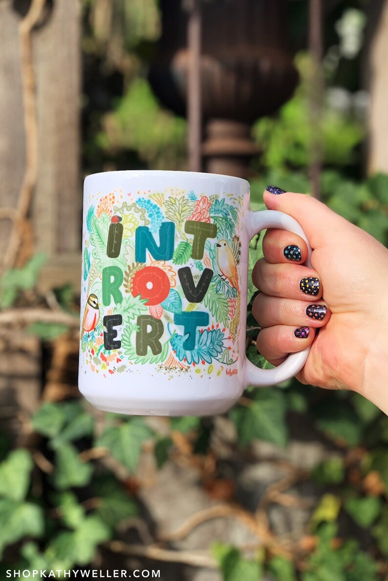 May include: White ceramic mug with a colorful floral design and the word "Introvert" written in a whimsical font. The mug is being held by a hand with black nail polish.