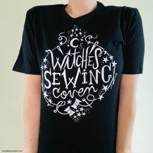 May include: Black t-shirt with a white graphic design that says "Witches Sewing Coven" surrounded by stars and sewing-related imagery.