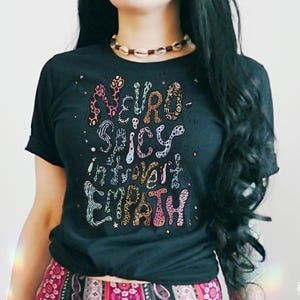 Könnte beinhalten: Ein schwarzes T-Shirt mit einem bunten Grafikdesign, auf dem "Neuro-Spicy Intuitive Empath" steht.