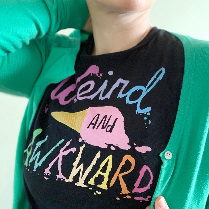 May include: A black t-shirt with the text "weird and awkward" in rainbow colors. The shirt features a pink and white ice cream cone graphic.