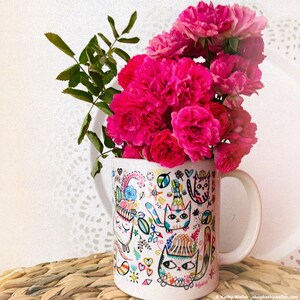 May include: A white ceramic mug with a colorful illustration of cats and other whimsical designs. The mug is filled with a bouquet of pink roses.