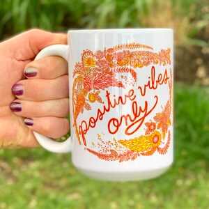 May include: White ceramic mug with a floral design in shades of orange and yellow. The mug has the text "positive vibes only" written in red cursive.