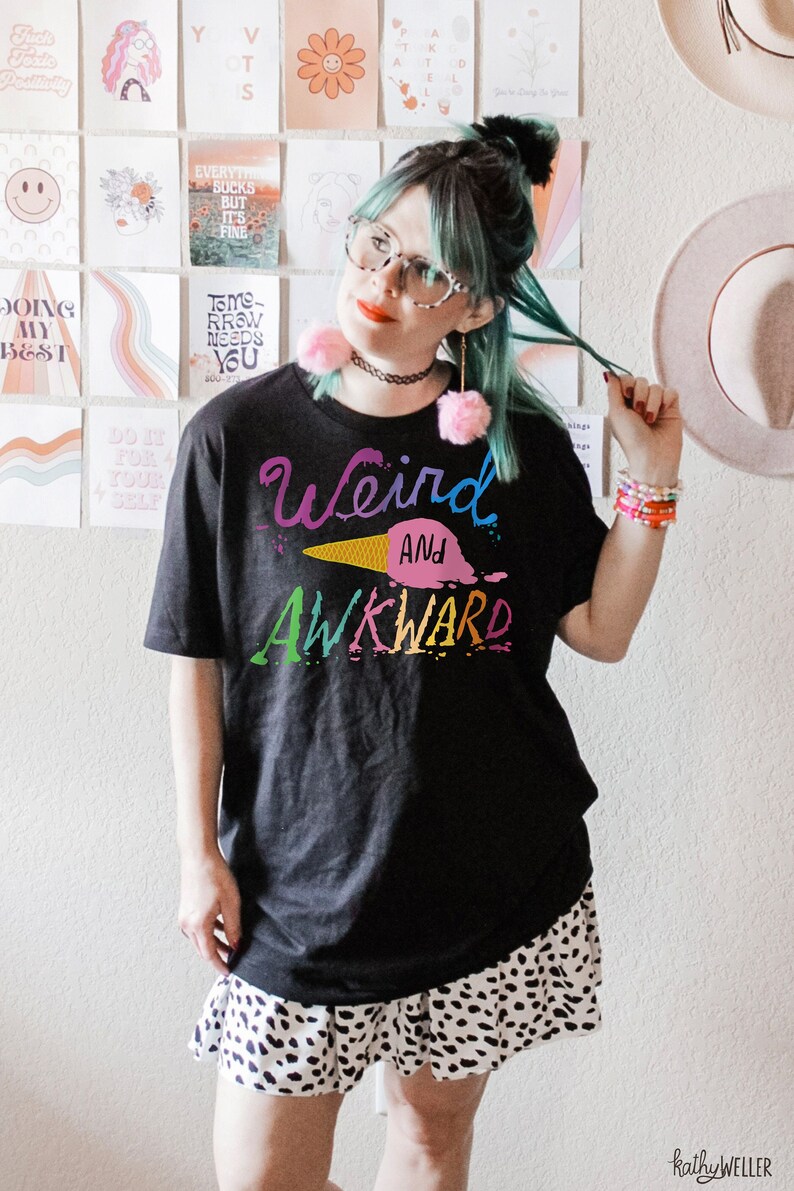 May include: A black t-shirt with the text "Weird and Awkward" in rainbow colors. The text is stylized with a dripping effect and includes an ice cream cone graphic. The person wearing the shirt is also wearing a black choker necklace and a white and black polka dot skirt.