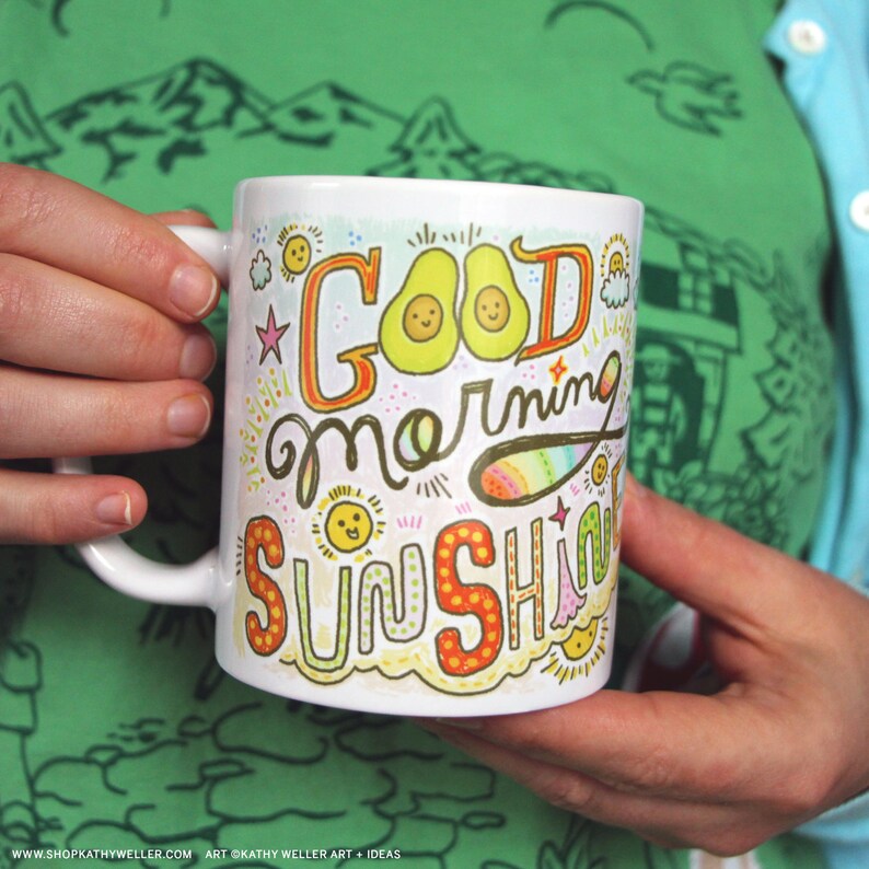 May include: White ceramic mug with a colorful hand-drawn design that says "Good morning sunshine" with two avocado illustrations. The design features yellow, orange, pink, green, and blue colors.