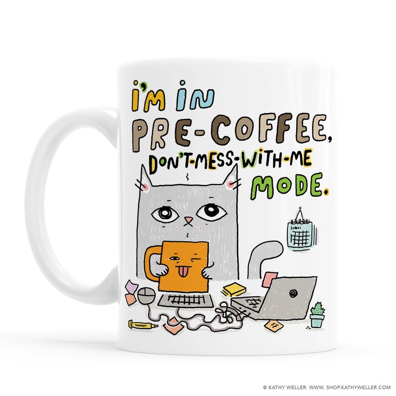 以下が含まれることがあります： 白いセラミック製のマグカップに、コーヒーカップを持った漫画の猫が描かれています。マグカップには「私はコーヒー前のモードです、邪魔しないでください。」という文字が書かれています。