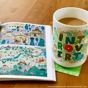 May include: A white coffee mug with a colorful floral design and the word "Introvert" printed in green letters. The mug is sitting on a green coaster and next to an open book with colorful illustrations.