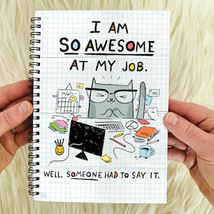 May include: A spiral bound notebook with a white background and a grid pattern. The notebook has a cartoon illustration of a cat wearing glasses and sitting at a desk with a computer, papers, and other office supplies. The text on the notebook reads "I am so awesome at my job. Well, someone had to say it."