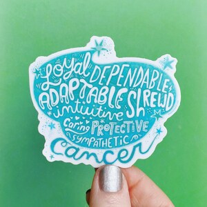 May include: A white sticker with a teal blue border and text. The sticker features the word "Cancer" in cursive, along with words like "Loyal," "Dependable," and "Adaptable." The background is a solid green.