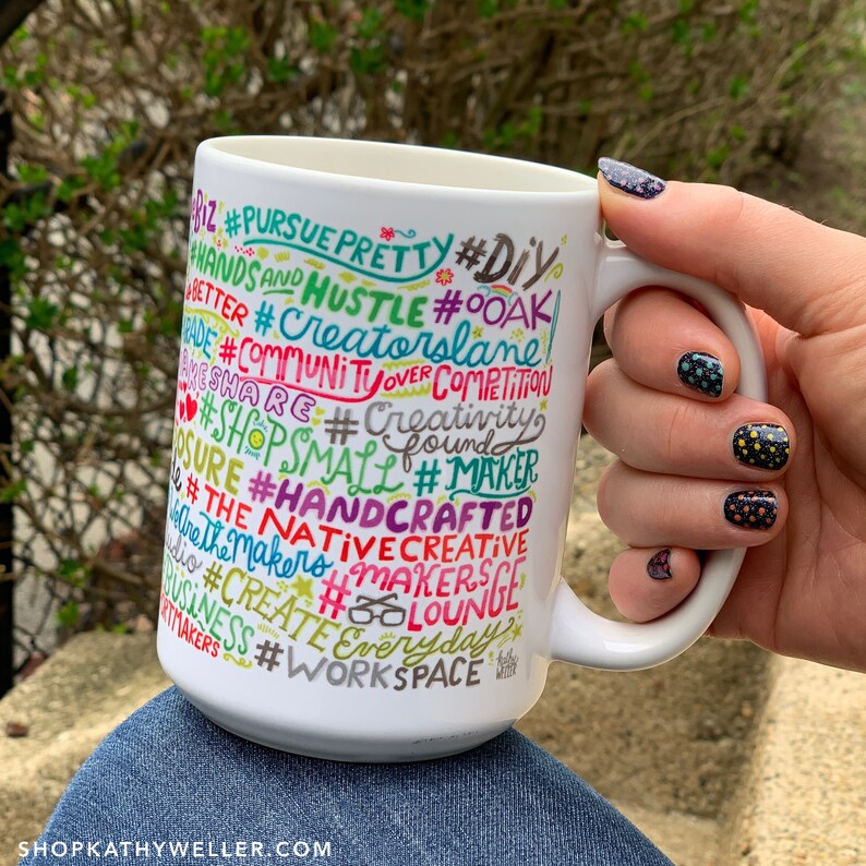 May include: White ceramic mug with a colourful design featuring the words "#PURSUE PRETTY #DIY #HANDS AND HUSTLE #OOAK #CREATORSLANE #COMMUNITUOVER COMPETITION #SHARE #CREATIVITY #SHOPSMALL #MAKER #HANDCRAFTED #THE NATIVECREATIVE #MAKERS #MAKERSLOUNGE #CREATE EVERYDAY #WORKSPACE"