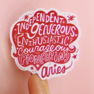 May include: A white sticker with a red heart shape and the words "Independent, Generous, Enthusiastic, Courageous, Pioneering" written in a pink and red font. The word "Aries" is written below the heart in a pink font.