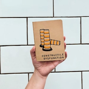 May include: A brown notebook with a black and white illustration of stacked orange and white construction cones. The text "CONSTRUCTILE DYSFUNCTION" is printed in black on the front cover.