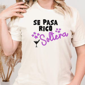 Op de afbeelding: Crèmekleurig T-shirt met de tekst "SE PASA RICO Soltera" in zwart en paars. Het ontwerp bevat een cocktailglas en sterren. Het is een casual kledingstuk.