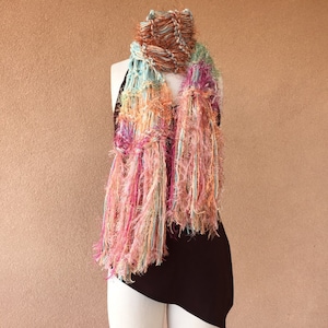 May include: A colorful, fringed scarf with shades of pink, orange, blue, and brown. The scarf is draped over a mannequin's neck.