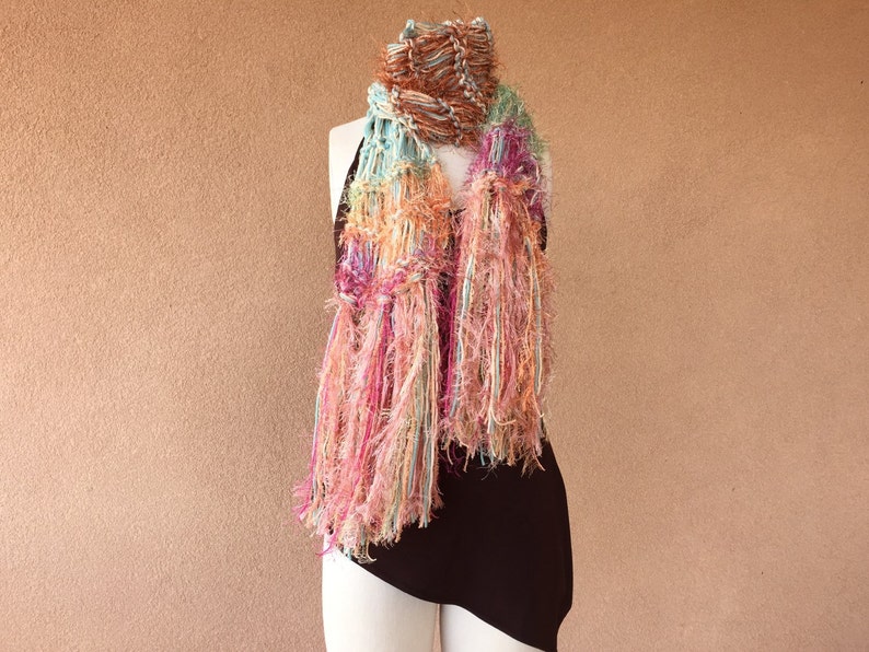 May include: A colorful, fringed scarf with shades of pink, orange, blue, and brown. The scarf is draped over a mannequin's neck.