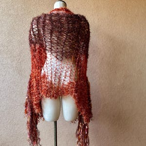 May include: A brown and orange knitted shawl with a fringe border. The shawl is draped over a mannequin.
