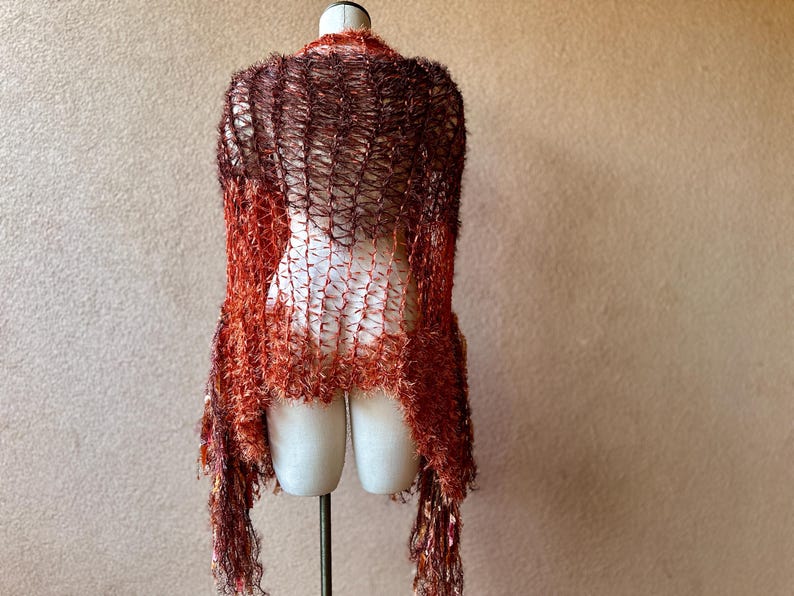 May include: A brown and orange knitted shawl with a fringe border. The shawl is draped over a mannequin.
