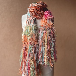 May include: A colorful scarf with long, fringed ends. The scarf is made of a variety of yarns in shades of orange, pink, blue, and green.