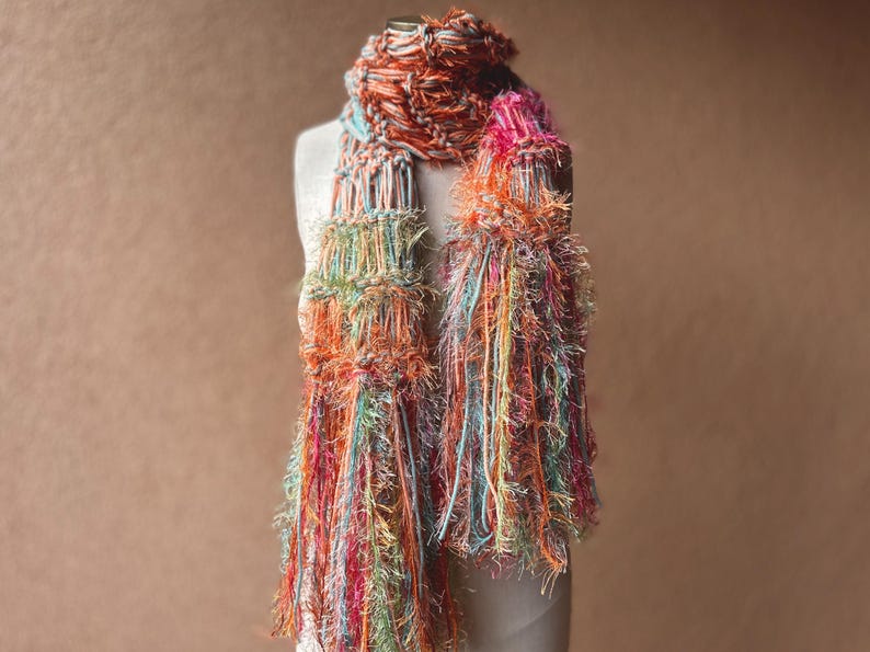 May include: A colorful scarf with long, fringed ends. The scarf is made of a variety of yarns in shades of orange, pink, blue, and green.