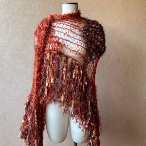 May include: A rust-colored crocheted shawl with fringe. The shawl is made of a soft, fuzzy yarn and has a delicate, openwork design.