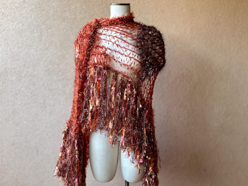 May include: A rust-colored crocheted shawl with fringe. The shawl is made of a soft, fuzzy yarn and has a delicate, openwork design.