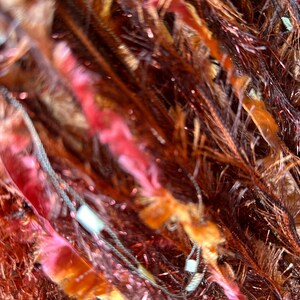 May include: Close-up of richly textured yarn fringe  in shades of deep red, burnt orange, and copper.  The fibers are long and slightly fuzzy, creating a sense of depth and movement.  Metallic threads are subtly interwoven, adding shimmer and visual interest.  They add a luxurious, multi-toned effect.
